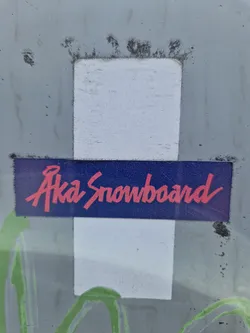A worn Aka Snowboard sticker on a surface. The sticker is mostly white with a dark blue band in the middle that contains the text Aka Snowboard in pink cursive lettering. 