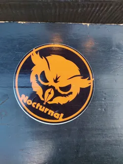 A round sticker featuring an orange owl design on a black background. The word 'Nocturnal' is written in orange below the owl. The sticker appears to be applied to a dark-colored surface.