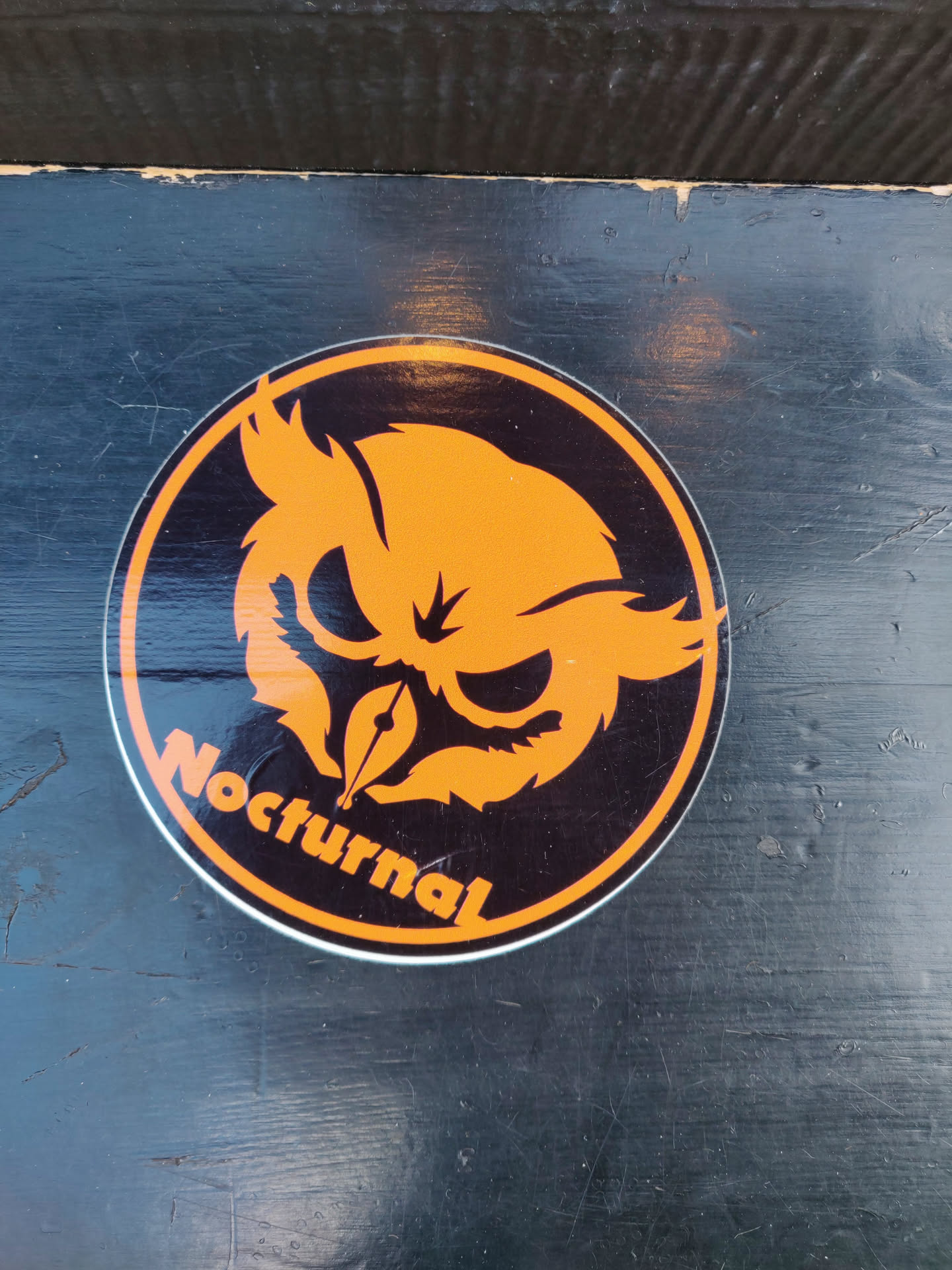 A round sticker featuring an orange owl design on a black background. The word 'Nocturnal' is written in orange below the owl. The sticker appears to be applied to a dark-colored surface.