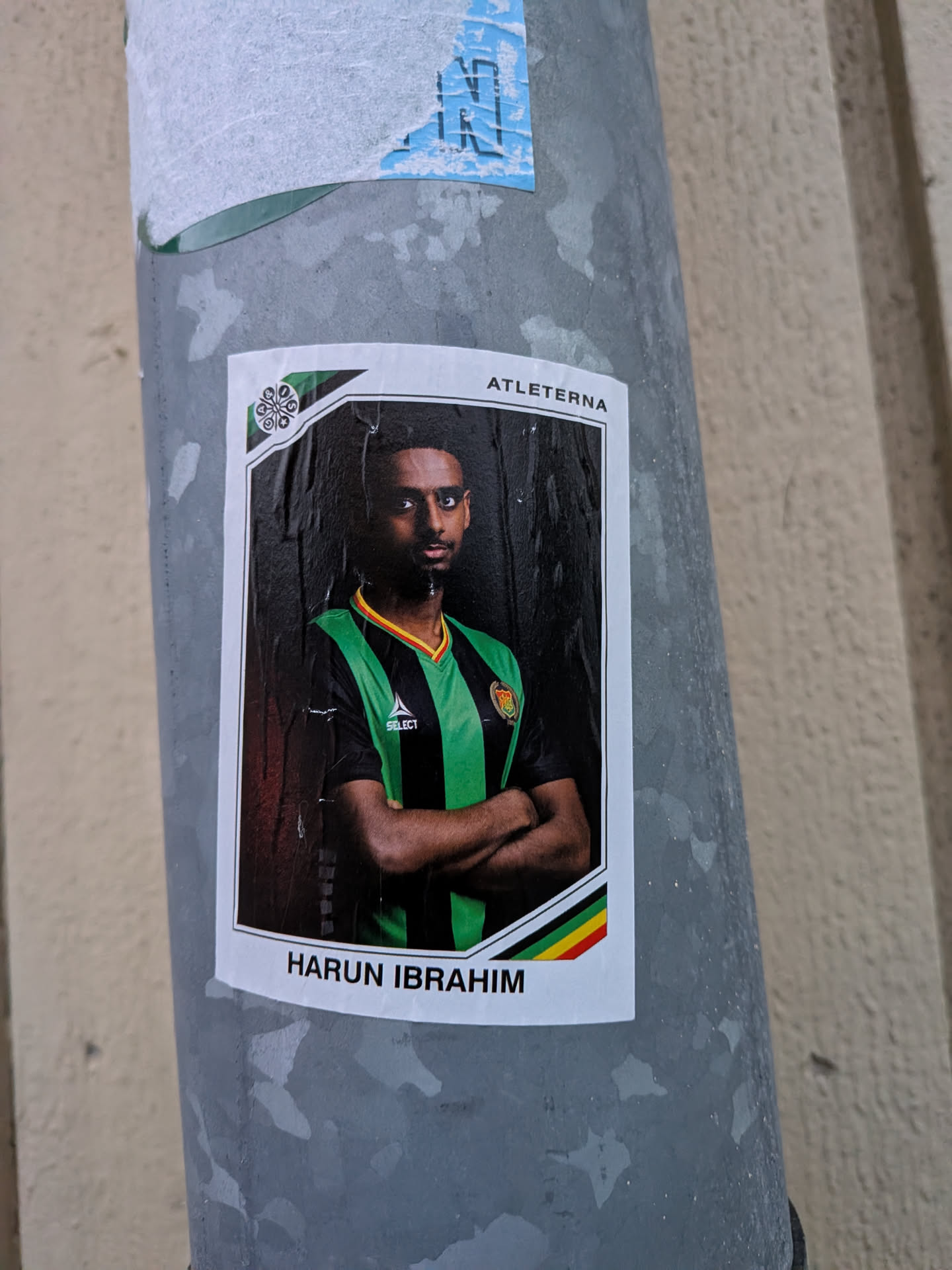 The sticker features a portrait of a man, presumably a footballer, named Harun Ibrahim. He is wearing a green and black striped jersey with a yellow and red trim around the collar. The jersey has a 'SELECT' logo and a crest on the left chest. The man has his arms crossed and is looking directly at the camera with a serious expression. The background is dark, creating a dramatic effect. The sticker has a white border with a green and black geometric design in the top left corner and a stripe of green, yellow, and red at the bottom. The text 'ATLETETERNA' is visible at the top, and 'HARUN IBRAHIM' is at the bottom.