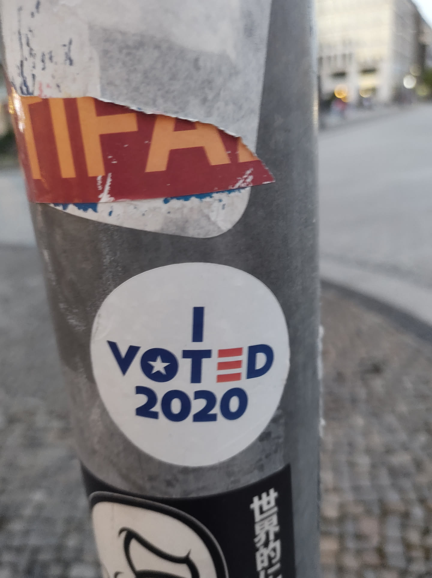 A white round sticker with blue text that says "I VOTED 2020".  There is a small star and three red stripes next to the word "VOTED". The sticker is affixed to a gray pole.