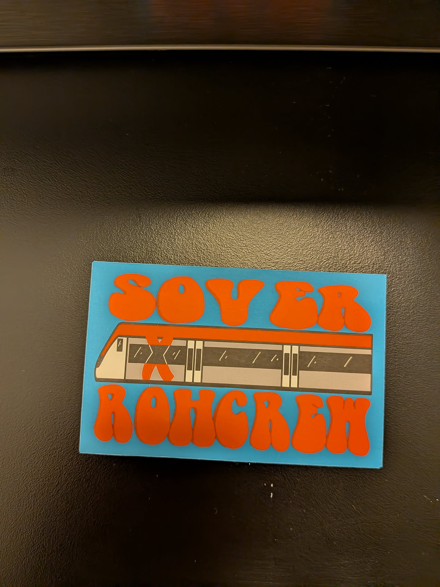 The sticker features a bright blue background. Centered on the sticker is a stylized illustration of a modern train or subway car, depicted in shades of white, gray, and black, with orange accents. Above the train, in large, bubbly, orange lettering, are the words 'SOV ER'. Below the train, also in large, bubbly, orange lettering, are the words 'ROHGRN'. The overall aesthetic is bold and graphic, with a retro feel.