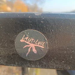 A round dark-colored sticker with a stylized creature and the text 'Laut + Luise' in a light pink/peach color. The creature resembles a simple, abstract insect or plant.