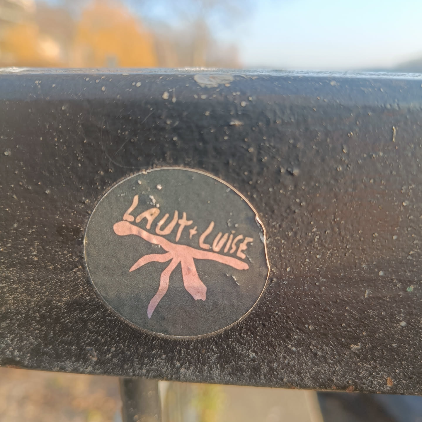 A round dark-colored sticker with a stylized creature and the text 'Laut + Luise' in a light pink/peach color. The creature resembles a simple, abstract insect or plant.