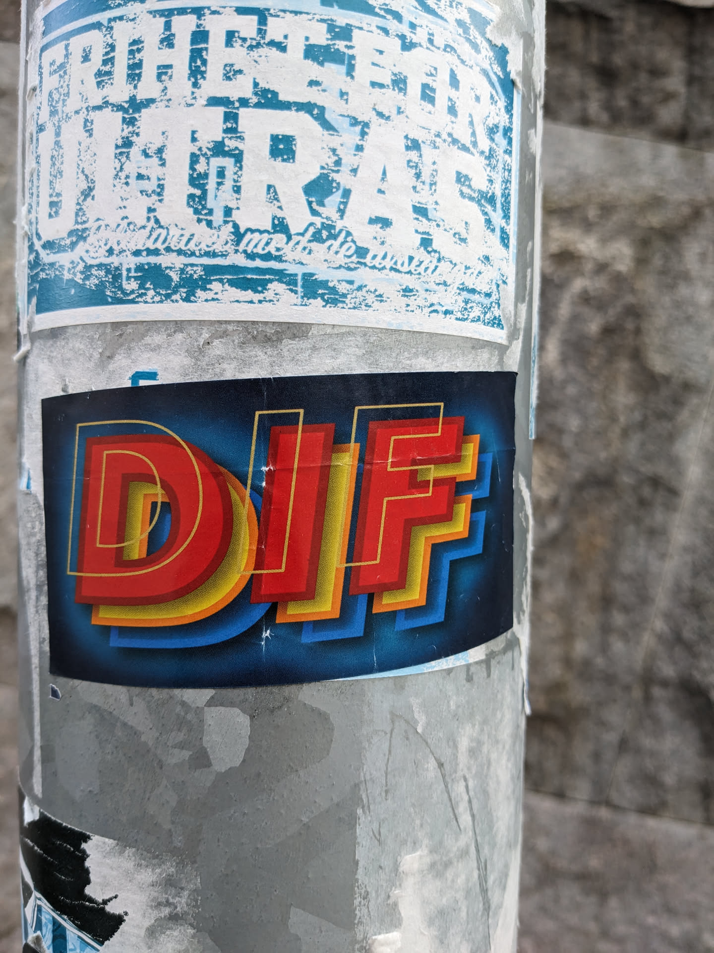 A sticker with the letters DIF in a retro style, with red, yellow, and blue colors. The background is dark blue. The sticker is attached to a grey pole.