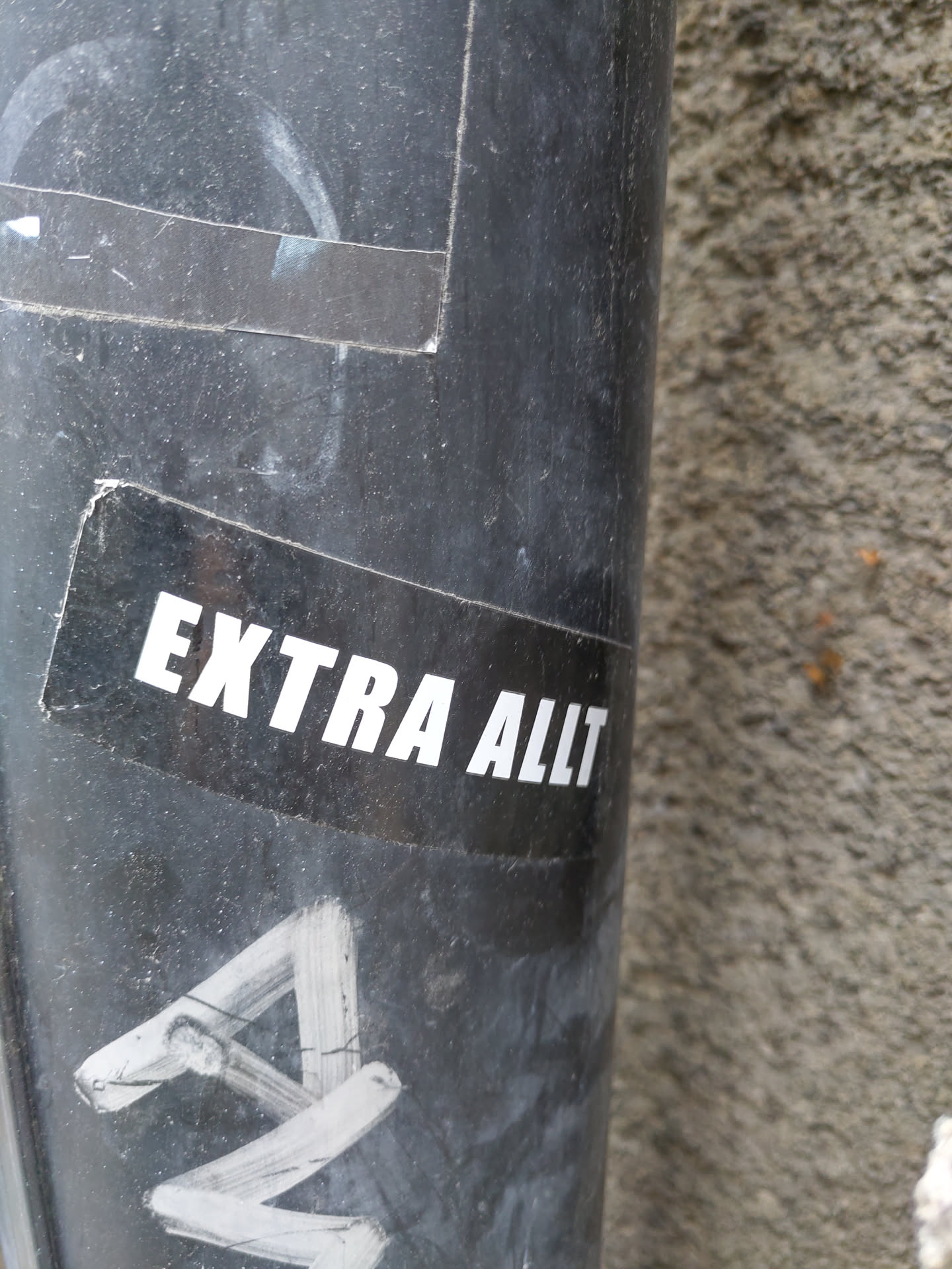 A black and white sticker with the text "EXTRA ALLT" in a bold, sans-serif font. The sticker is affixed to a dark-colored cylindrical surface.