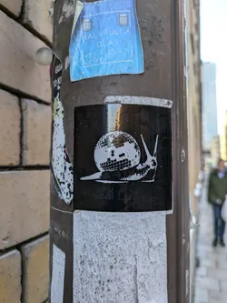 A black and white sticker of a snail carrying a disco ball on its back. The sticker is affixed to a brown pole.
