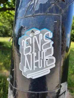 A silver and light blue sticker with stylized text that appears to say 'SNS NHB'. The sticker is applied to a dark-colored pole outdoors. The style is reminiscent of street or graphic art, with a slightly 3D effect to the lettering.
