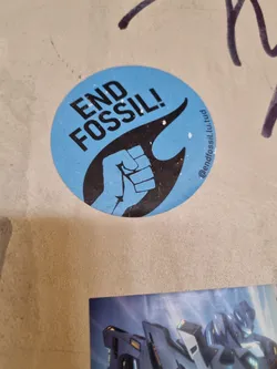 A round sticker with a clenched fist in a flame and the words "END FOSSIL!" on a light blue background.  The sticker also includes a social media handle.