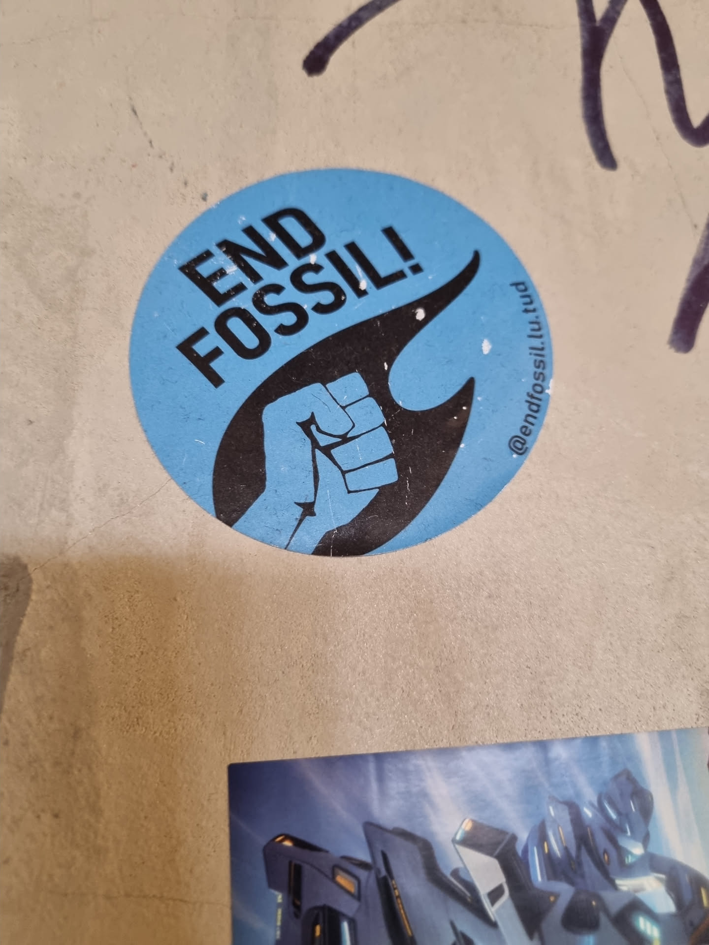 A round sticker with a clenched fist in a flame and the words "END FOSSIL!" on a light blue background.  The sticker also includes a social media handle.