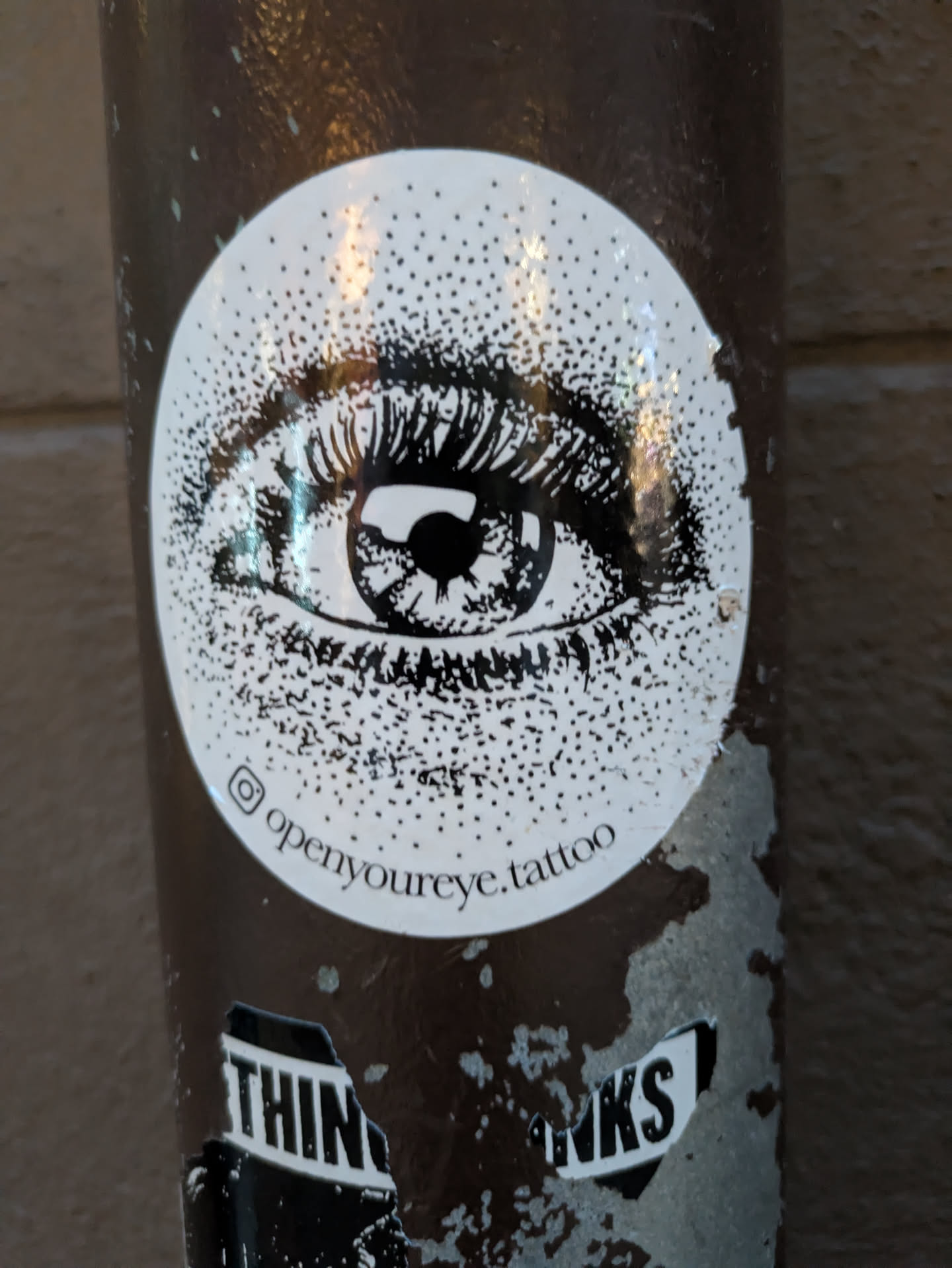 The sticker is circular and predominantly white with black detailing. At its center is a detailed illustration of a human eye, focusing on the iris and eyelashes. The eye is rendered in black ink, with the iris showing concentric rings and a dark pupil. The eyelashes are sharp and expressive. The white background of the sticker is filled with a dense pattern of black dots, creating a textured or stippled effect that extends from the eye outwards towards the edges of the circle. Below the eye illustration, curved text is visible along the bottom edge of the sticker.