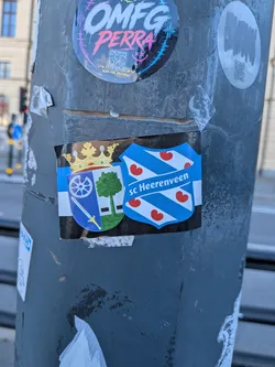 Street sticker A rectangular sticker placed horizontally on a metal street pole. It displays the traditional coat of arms of Heerenveen, crowned in gold with a wheel, sword, and tree, next to the shield crest of Dutch football club sc Heerenveen featuring blue and white diagonal stripes adorned with red water-lily leaves (pompebêden). The background features horizontal stripes in blue, white, and black.