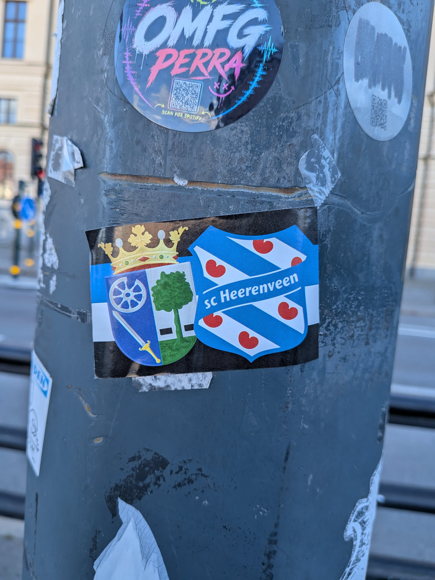 A rectangular sticker placed horizontally on a metal street pole. It displays the traditional coat of arms of Heerenveen, crowned in gold with a wheel, sword, and tree, next to the shield crest of Dutch football club sc Heerenveen featuring blue and white diagonal stripes adorned with red water-lily leaves (pompebêden). The background features horizontal stripes in blue, white, and black.