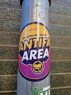 A round sticker with a purple and yellow design.  It features the text "Antifa Area" prominently, along with the slogans "No Cops! No Nazis!" and "No borders". A symbol resembling a circle with a flag inside is also visible. The sticker appears to be affixed to a metal pole.