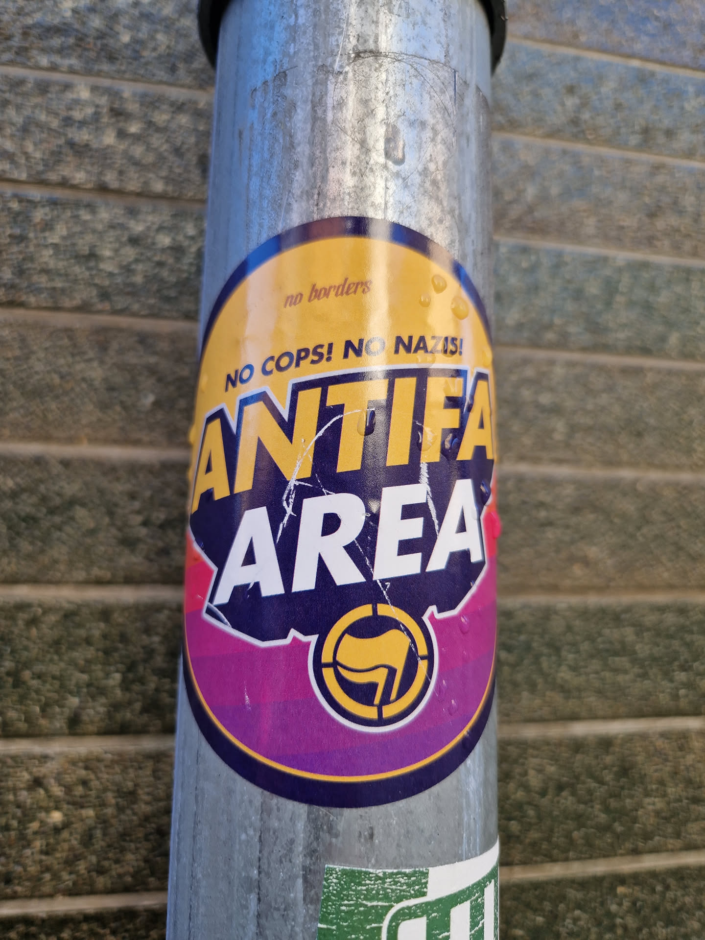 A round sticker with a purple and yellow design.  It features the text "Antifa Area" prominently, along with the slogans "No Cops! No Nazis!" and "No borders". A symbol resembling a circle with a flag inside is also visible. The sticker appears to be affixed to a metal pole.