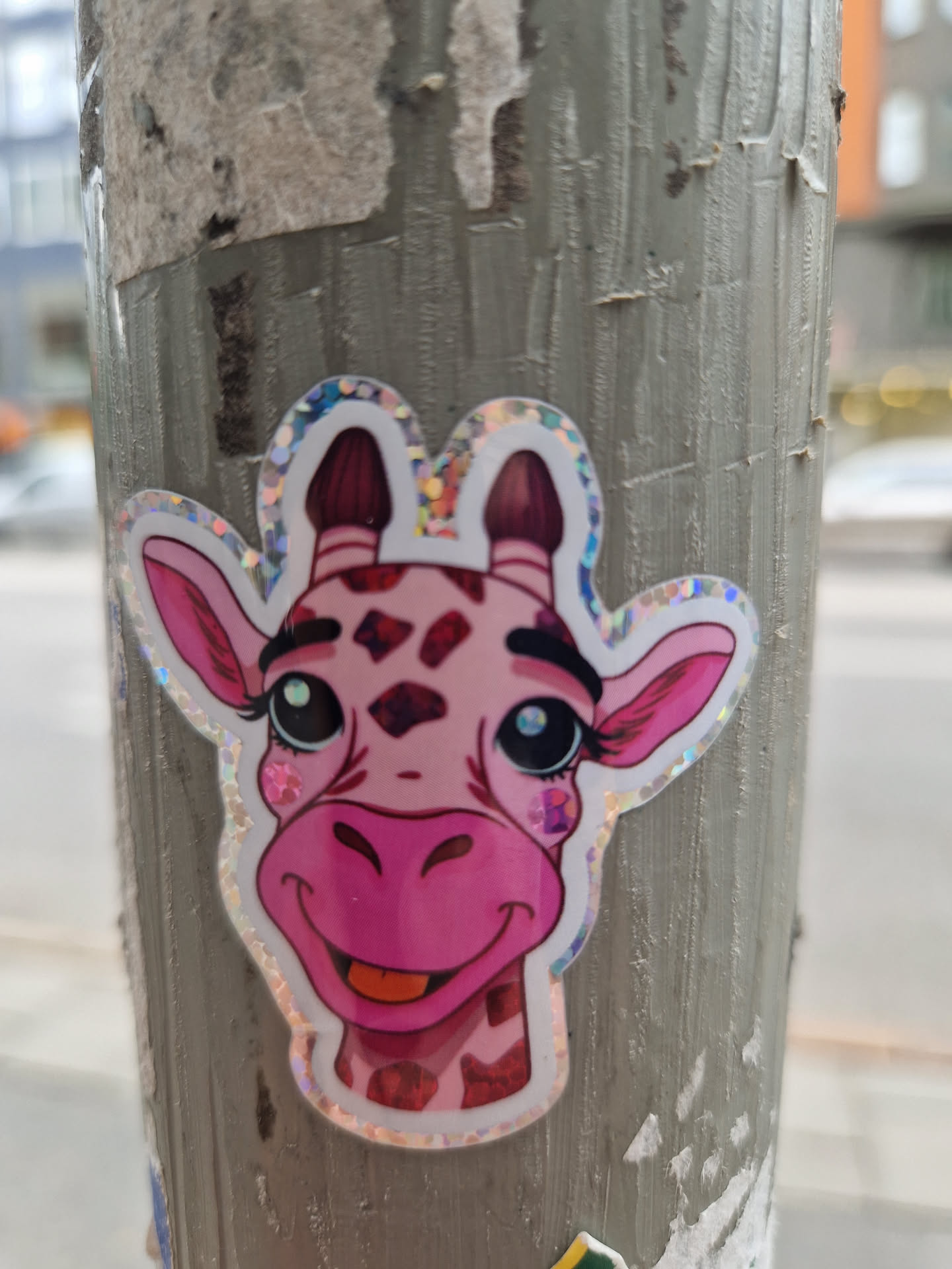 A cute pink giraffe sticker with sparkly accents, plastered on a pole.  The giraffe has big eyes and a happy expression. 