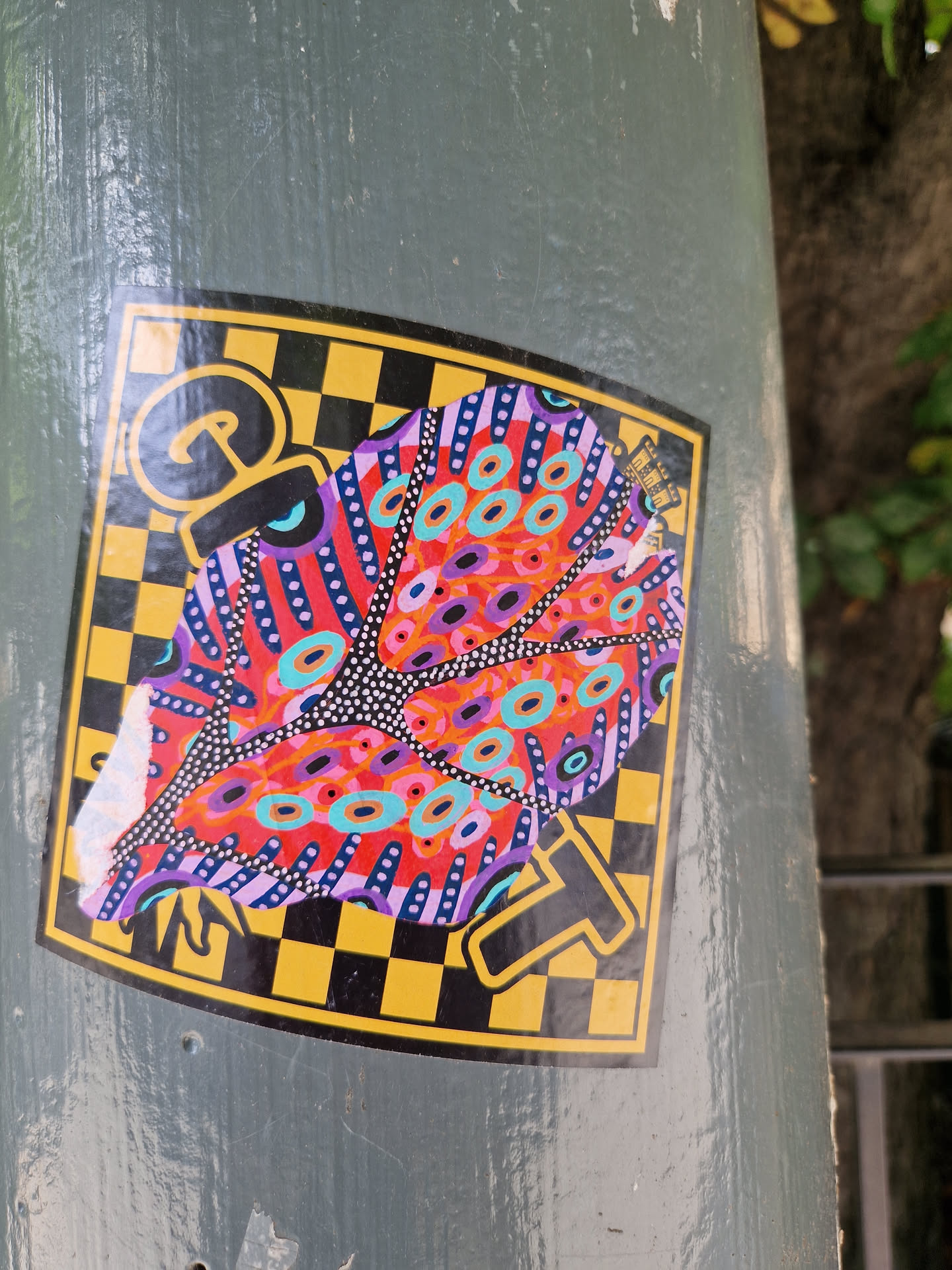 A colorful sticker featuring an abstract design resembling a butterfly on a checkered background. The main colors are red, pink, purple, turquoise, and black on a yellow checkered background.  A partially visible letter 'G' and 'T' are present in the corners. The sticker has a slightly damaged corner.