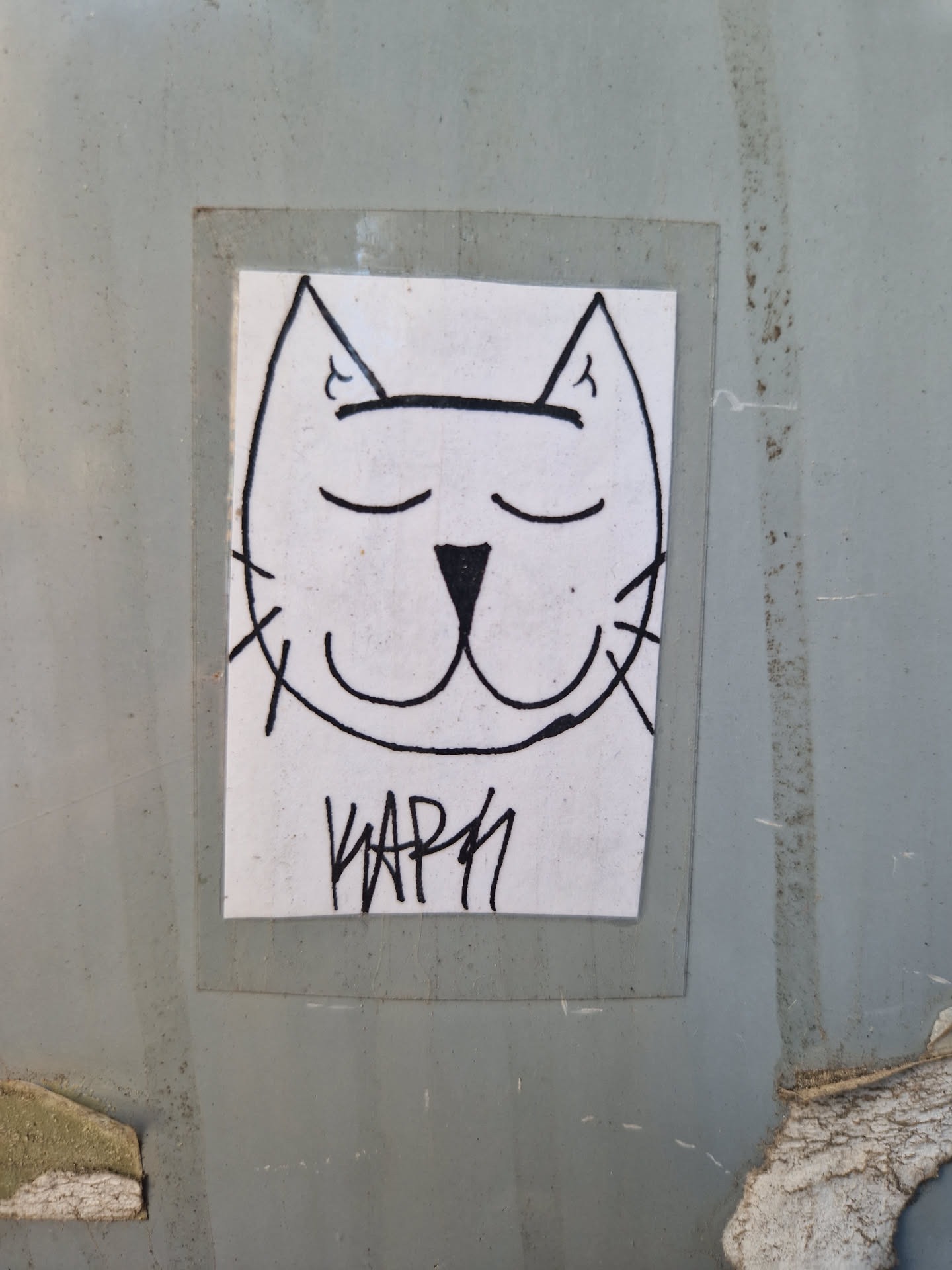 A sticker featuring a minimalist line art drawing of a cat's face. The cat has its eyes closed and a slight smile. The artist's signature, "KAPN", is written beneath the drawing.