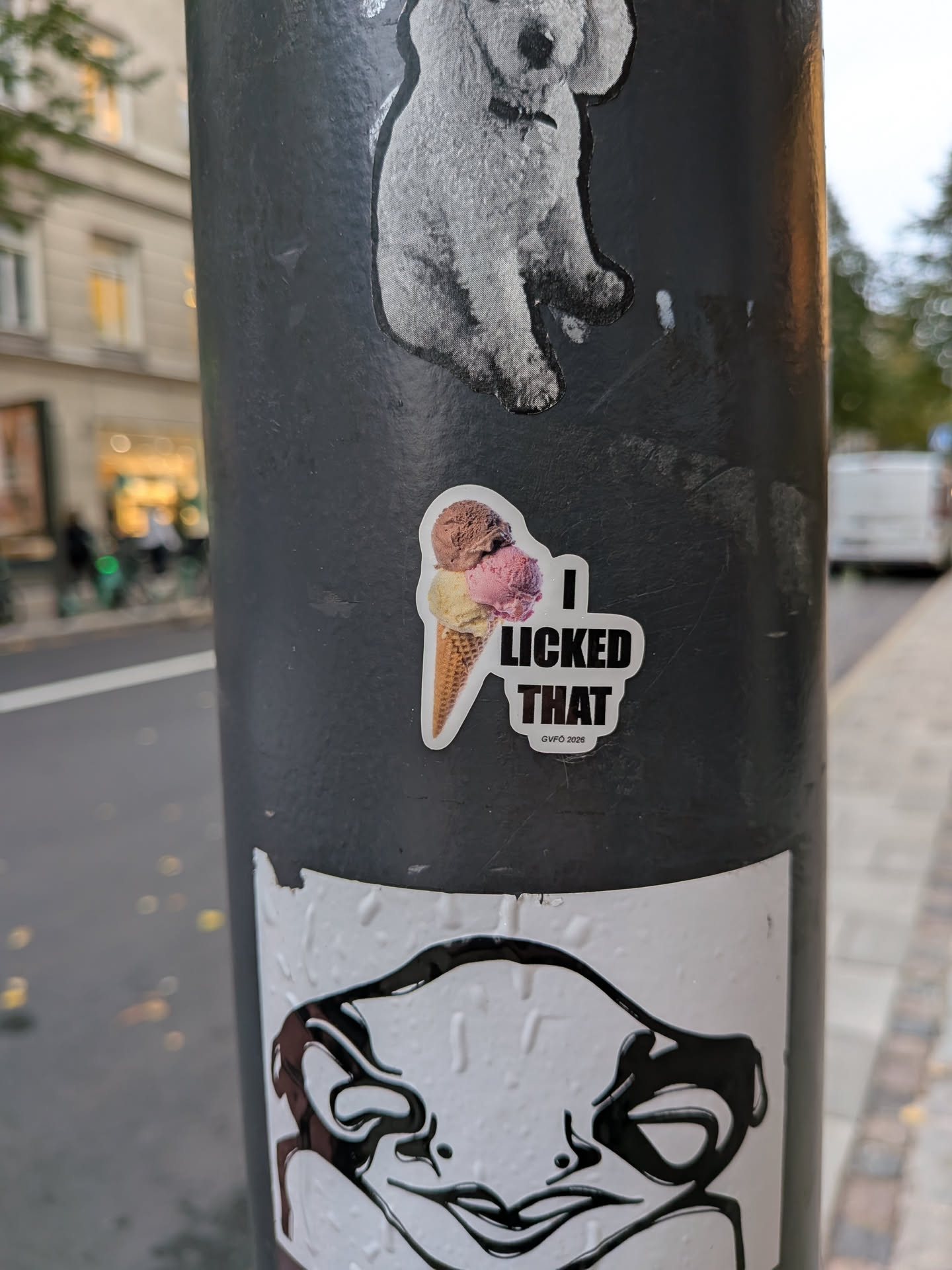 A die-cut sticker placed on a dark pole featuring a realistic image of an ice cream waffle cone with three scoops of ice cream: chocolate, strawberry, and vanilla. Next to the cone, bold black text reads 'I LICKED THAT' set against a white background contour, with smaller text underneath saying 'GVFÖ 2026'.