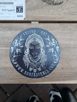 Round black and white sticker with an illustration of a deity-like figure with intricate details, surrounded by floral elements and skulls at the bottom. The text "Tattoo Art" is visible at the top and the website address "www.ronyroyart.com" is visible at the bottom.