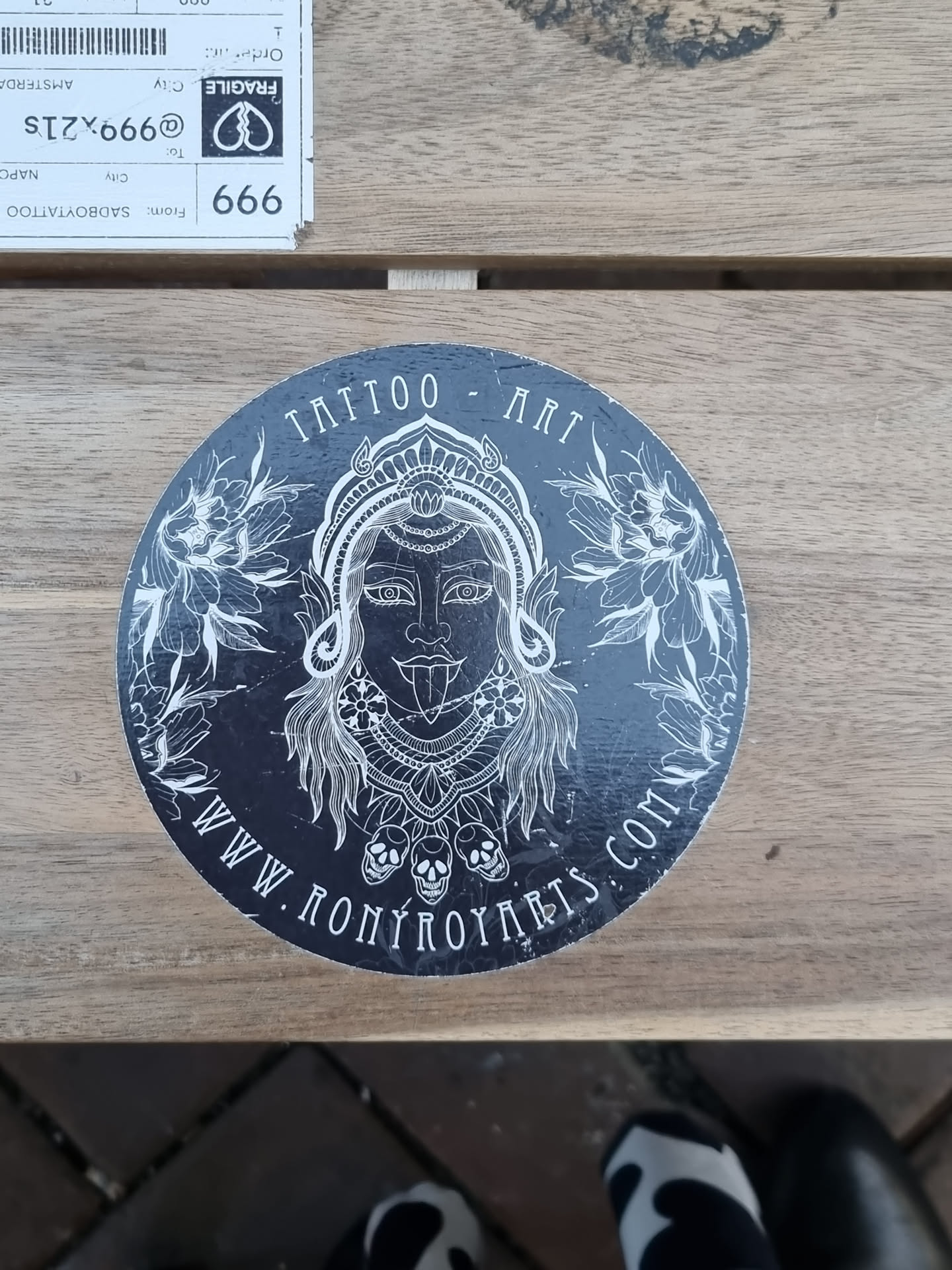 Round black and white sticker with an illustration of a deity-like figure with intricate details, surrounded by floral elements and skulls at the bottom. The text "Tattoo Art" is visible at the top and the website address "www.ronyroyart.com" is visible at the bottom.