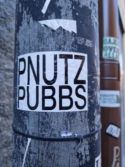 A black and white sticker with the text PNUTZ PUBBS on a weathered pole. 