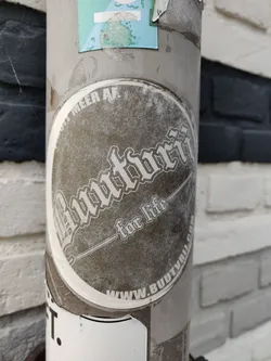 A round, weathered sticker with the text "Buutvrij" in a stylized font. Below it, it says "for life." At the bottom, there's a website address: www.buutvrij.com