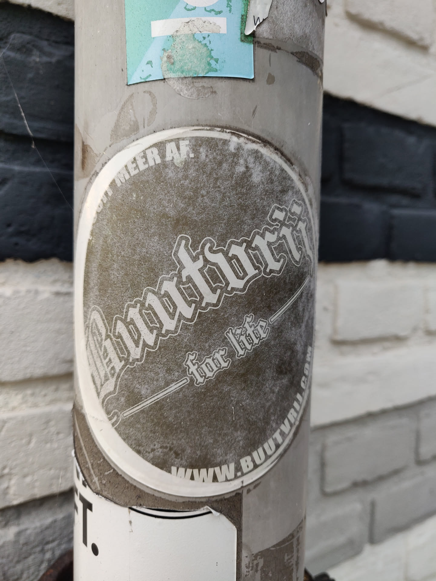 A round, weathered sticker with the text "Buutvrij" in a stylized font. Below it, it says "for life." At the bottom, there's a website address: www.buutvrij.com