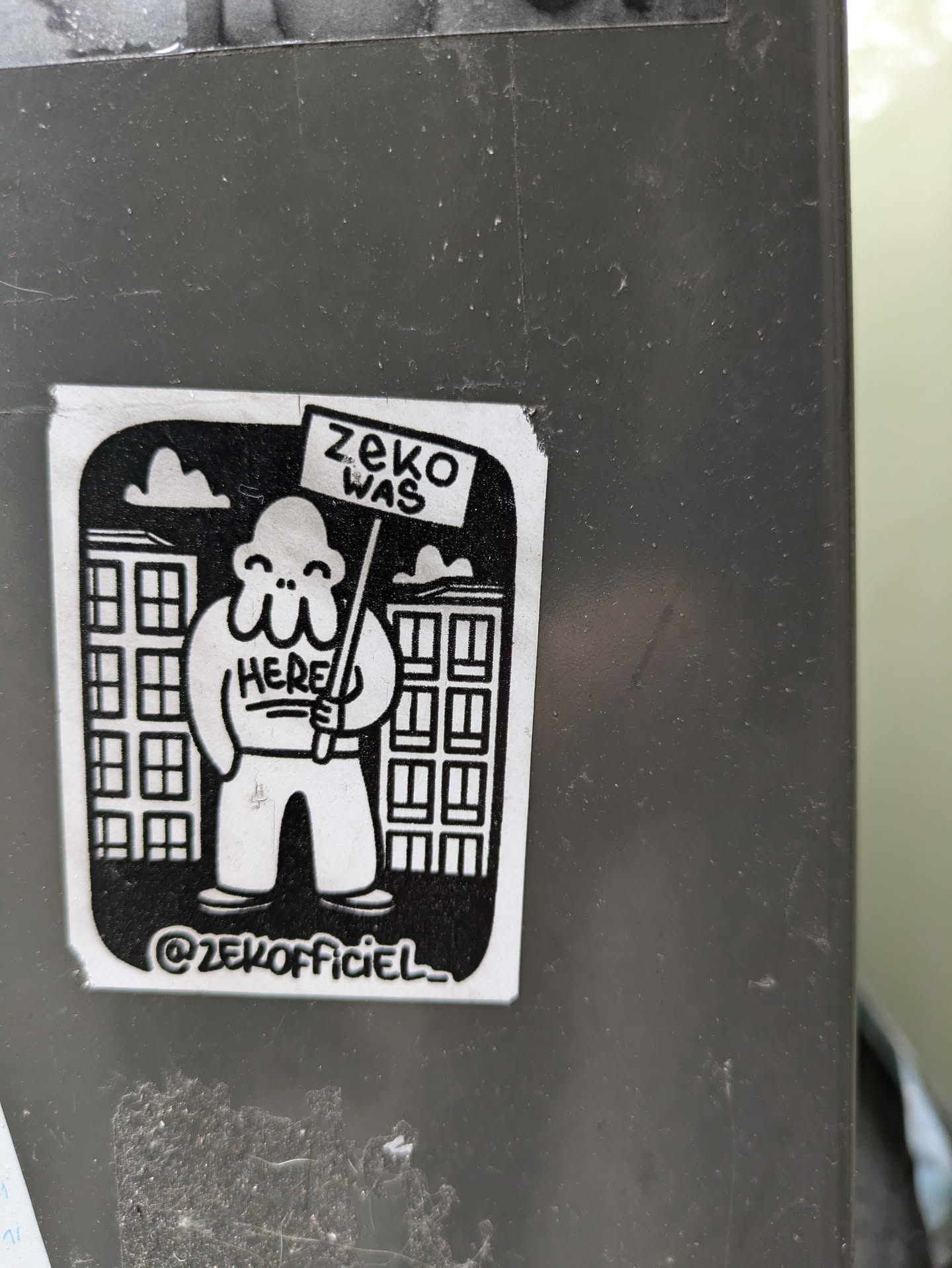 Black and white sticker featuring a cartoon character holding a sign that says "ZeKO WAS HERE". The character is standing in front of buildings. The sticker has a simple design and uses bold outlines. At the bottom, there's an Instagram handle: @zeKOfficiel_