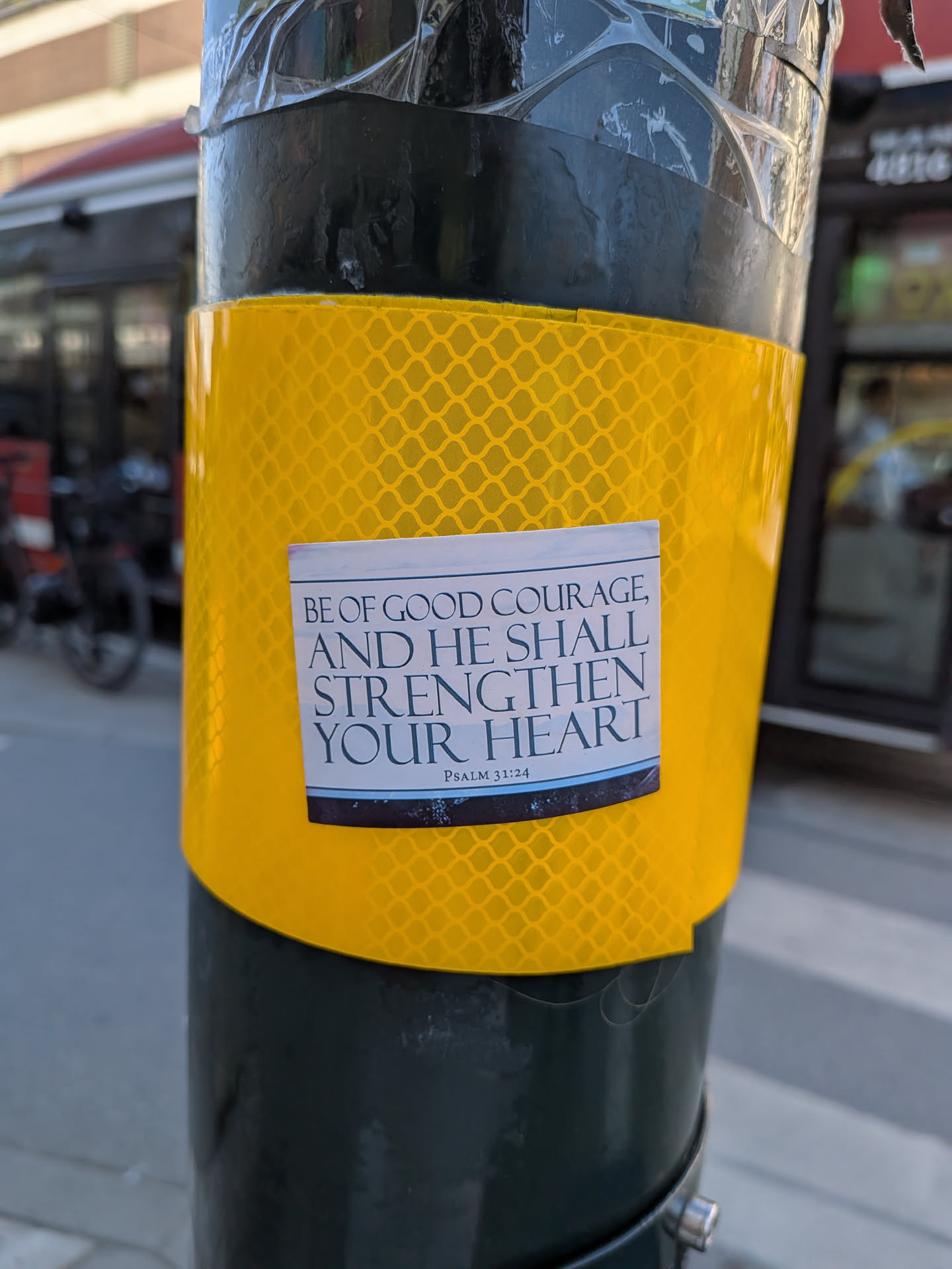 A rectangular sticker with a clean white background and black serif font. The main text reads "BE OF GOOD COURAGE, AND HE SHALL STRENGTHEN YOUR HEART". Below this, in a smaller font, it specifies "PSALM 31:24". The bottom edge of the sticker features a thin blue line above a dark, textured border that appears to be a mix of deep blue and black. The sticker is adhered to a bright yellow, reflective surface with a honeycomb pattern, which is wrapped around a dark grey or black pole.
