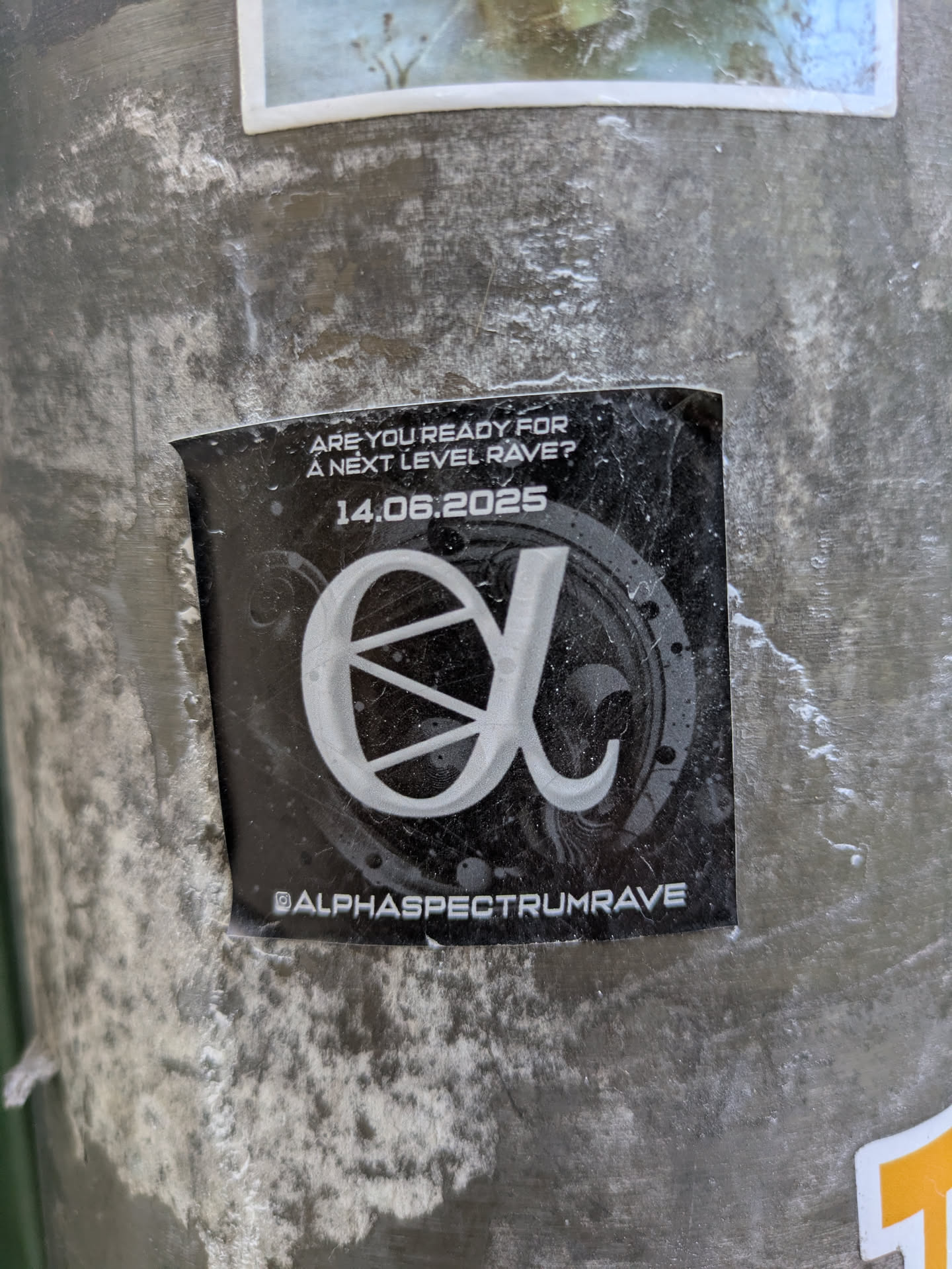 A black sticker advertising an upcoming event. It features the text "ARE YOU READY FOR A NEXT LEVEL RAVE?" and the date "14.06.2025" above a stylized logo resembling the Greek letter alpha within a speaker graphic. At the bottom, the social media handle @ALPHASPECTRUMRAVE is printed. The sticker is affixed to a weathered metal pole.
