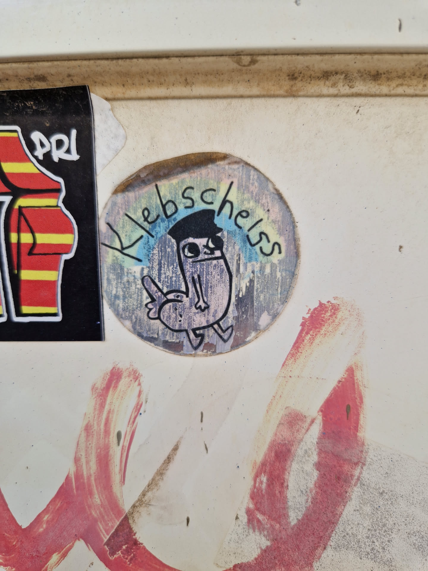 Round sticker with a cartoon drawing of a bird-like creature wearing a cap. The text "Klebscheiss" is written around the drawing. 
