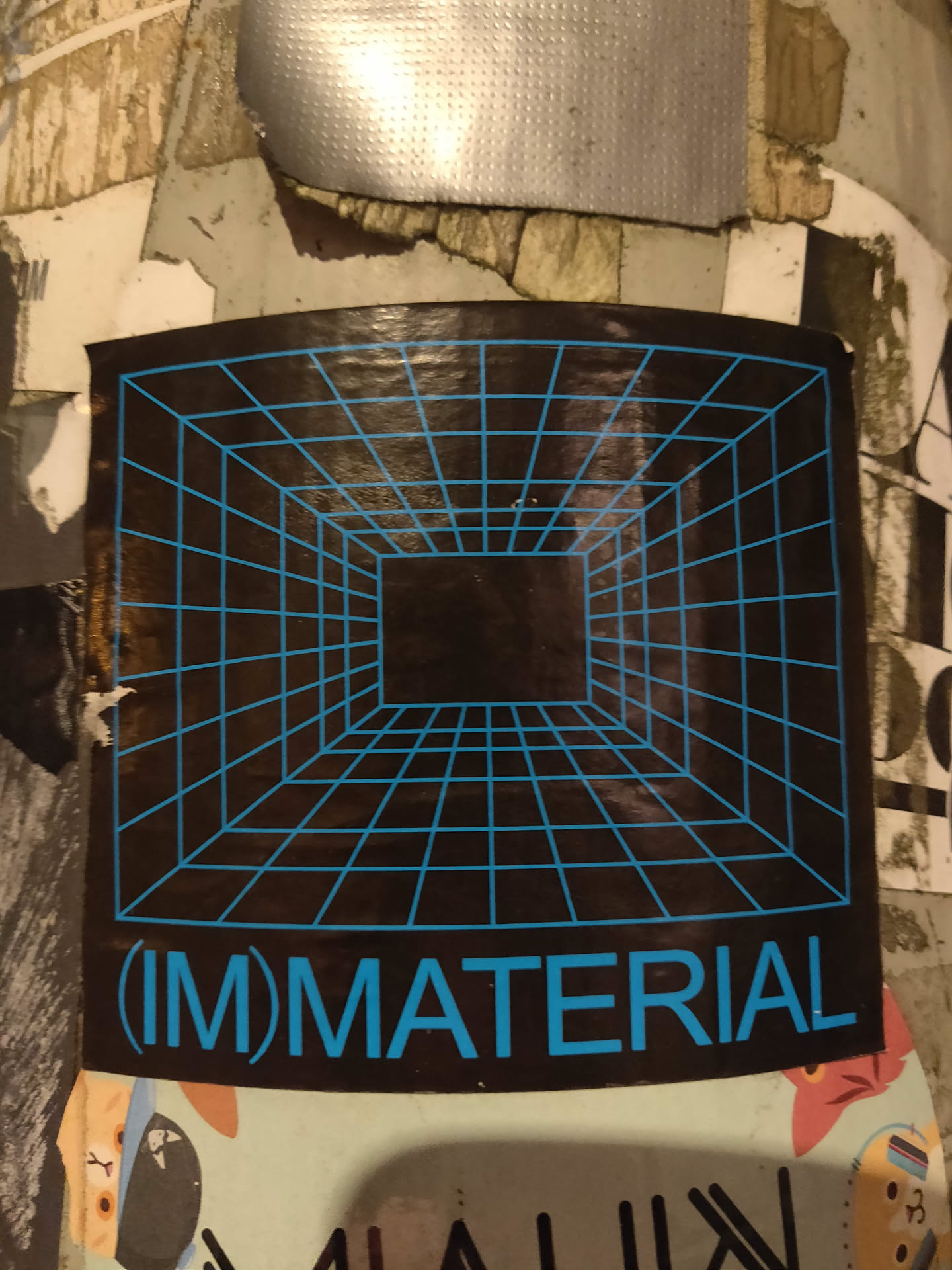 A dark sticker with a cyan grid forming a perspective cube and the text (IM)MATERIAL in cyan. 