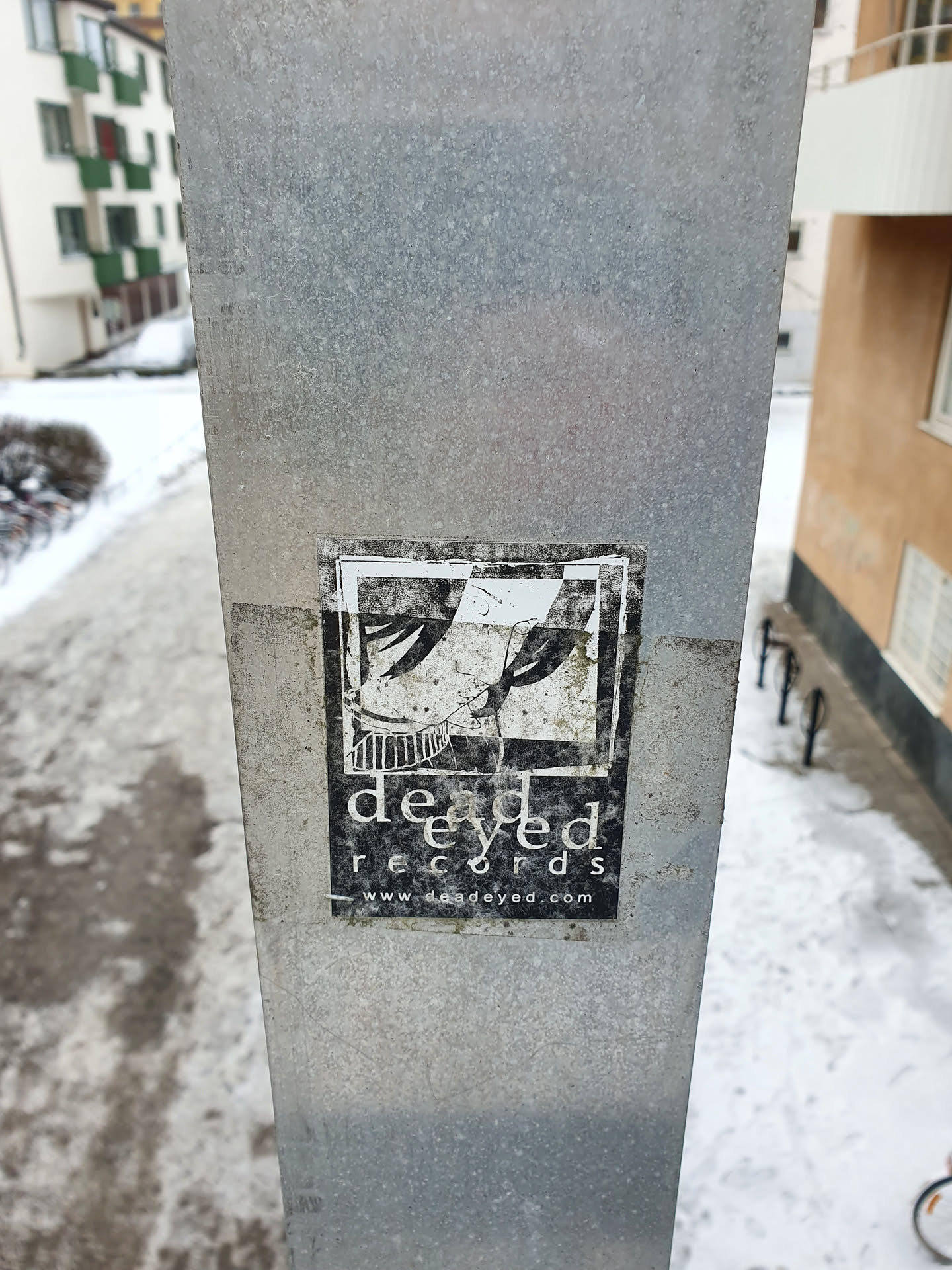 A black and white sticker featuring the Deadeye Records logo. The logo depicts a pair of legs and feet, possibly wearing boots, with a simple design.  Below the image, the text 'deadeye records' and a website address 'www.deadeyed.com' are visible. The sticker appears to be affixed to a metal pole outdoors, with some snow visible in the background.