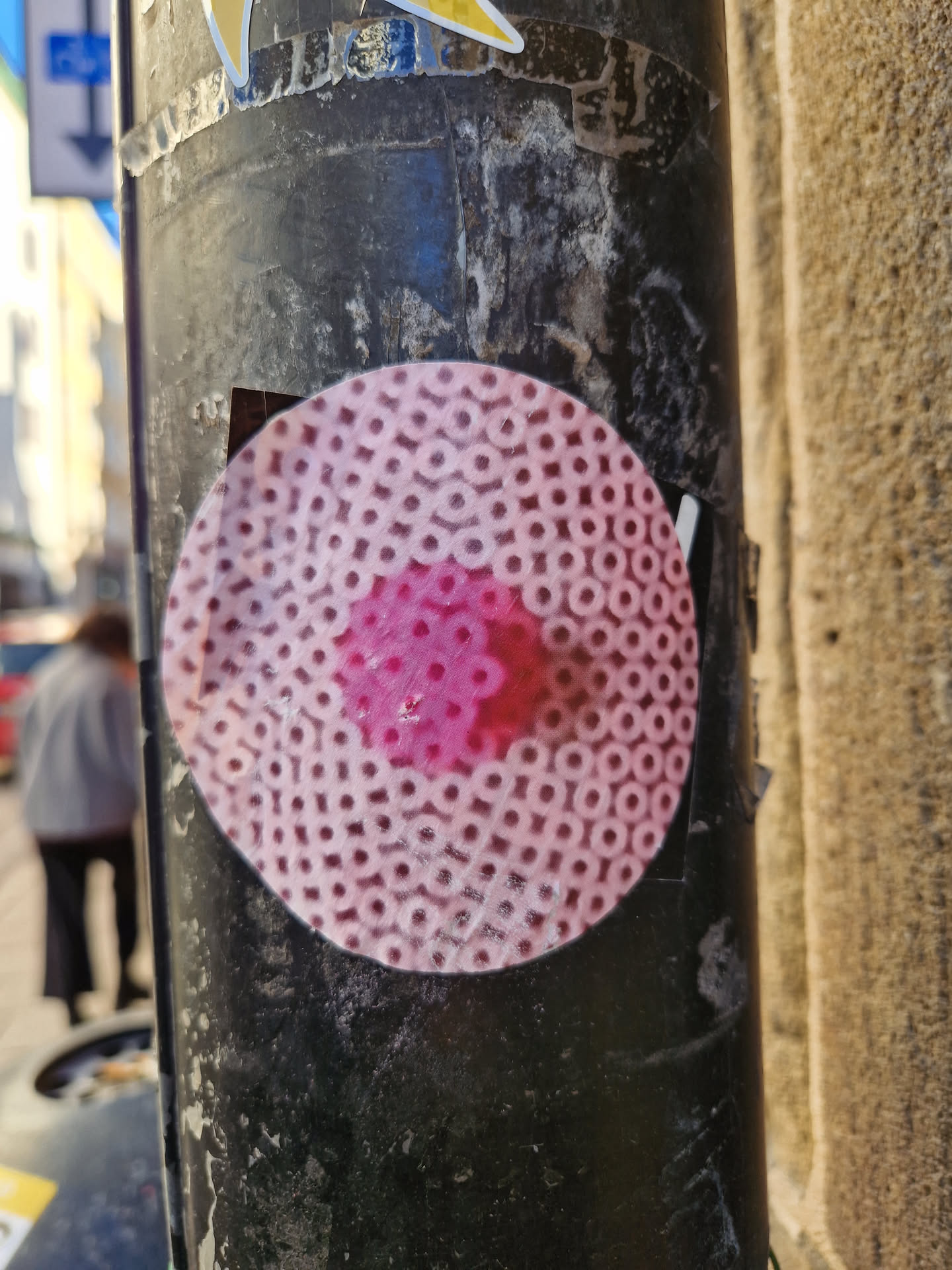 Round sticker featuring a pattern of small circles in pink and white. The central area is a darker pink. The sticker is affixed to a dark-colored pole, showing some wear and tear.