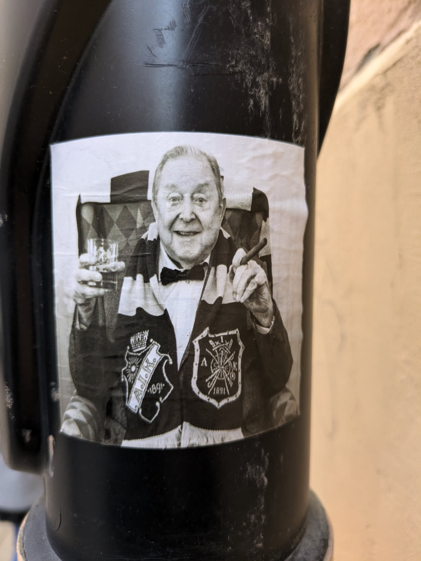 Black and white sticker featuring a photo of an older man with a cigar and a drink, wearing a sweater with embroidered patches. The photo has a vintage look and feel. The sticker is affixed to a dark pole.