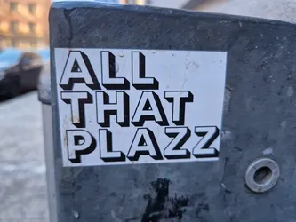A black and white sticker with the text "ALL THAT PLAZZ" in a stylized, three-dimensional font. The sticker is affixed to a gray metal surface.