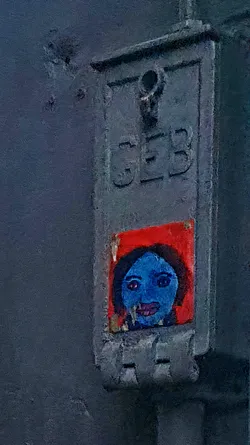 A sticker on a gray metal box features a portrait of a person with blue skin and a red background. The portrait is painted in a simple, almost childlike style, with large eyes and dark hair. The sticker is placed below the letters 'CEB' embossed into the metal.