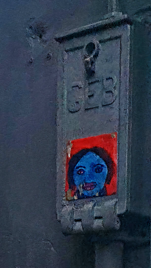 A sticker on a gray metal box features a portrait of a person with blue skin and a red background. The portrait is painted in a simple, almost childlike style, with large eyes and dark hair. The sticker is placed below the letters 'CEB' embossed into the metal.