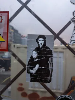 The sticker is a black and white photographic print, showing Greta Thunberg in the foreground, looking directly at the viewer with her arms crossed. Behind her, a figure wearing a black balaclava with eye and mouth holes stands, also with arms crossed. The sticker appears to be slightly creased and worn, indicating it has been applied to a surface. The overall impression is one of solidarity and a strong message of protest.