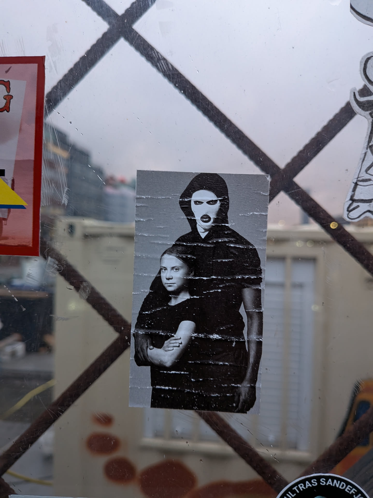 The sticker is a black and white photographic print, showing Greta Thunberg in the foreground, looking directly at the viewer with her arms crossed. Behind her, a figure wearing a black balaclava with eye and mouth holes stands, also with arms crossed. The sticker appears to be slightly creased and worn, indicating it has been applied to a surface. The overall impression is one of solidarity and a strong message of protest.