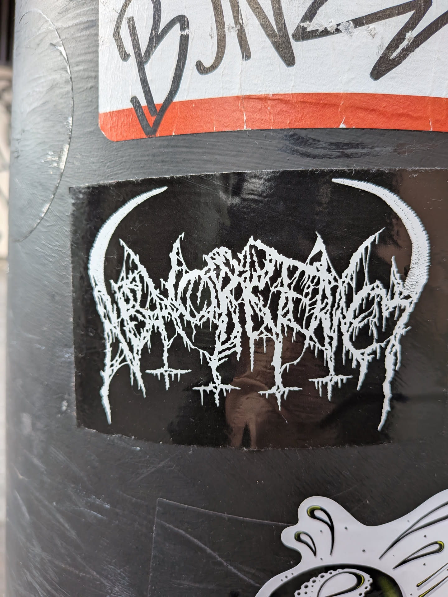 A rectangular black sticker featuring a highly stylized, intricate white logo at its center. The logo is rendered in a 'spiky' or 'dripping' font, characteristic of extreme metal genres, making it almost illegible. It is framed on both the left and right sides by prominent white crescent moon or horn-like shapes that curve inwards towards the logo. Below the main logo, there are several smaller, inverted cross-like or spike-like elements. The sticker has a slightly glossy finish, showing some reflections, and appears to be adhered to a dark, possibly metallic, pole.