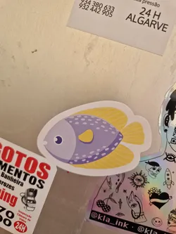 A sticker featuring a cartoon fish with purple body, yellow fins and tail, and white dots. It has a simple, cute design.