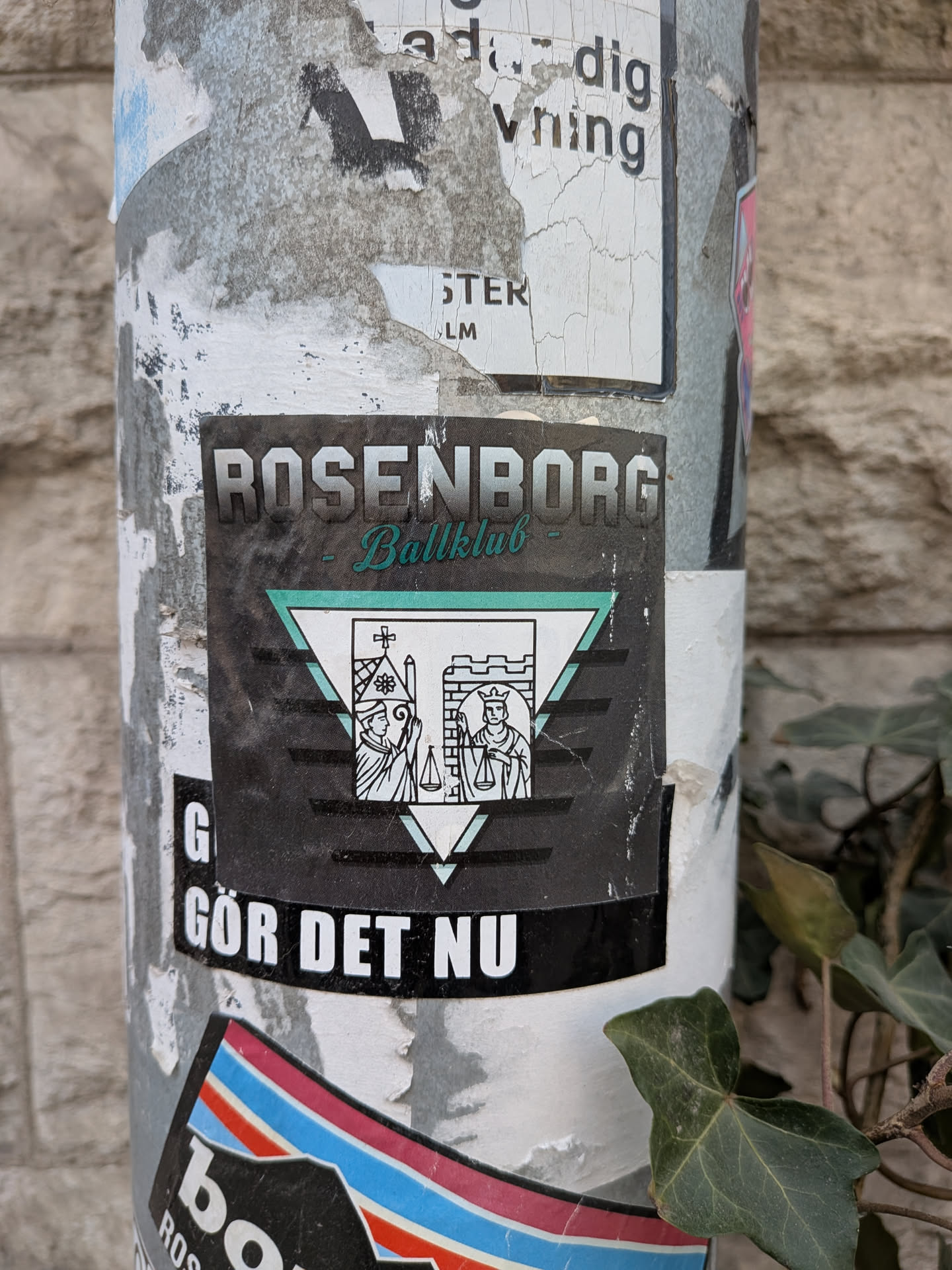 A rectangular sticker, predominantly black, featuring the name 'ROSENBORG' in bold, white, block letters at the top, with 'Ballklub' in a flowing, teal-green script directly beneath it. The central design is an inverted teal-green outlined triangle, within which a smaller, upright white triangle is framed. Inside this inner triangle, two figures are depicted in black line art: on the left, a cleric or bishop holding scales next to a church-like structure with a cross; on the right, a king or ruler also holding scales next to a castle tower. Below this emblem, the phrase 'GÖR DET NU' is written in bold, white, sans-serif capital letters. The sticker shows signs of wear and minor scuffs, consistent with outdoor exposure.