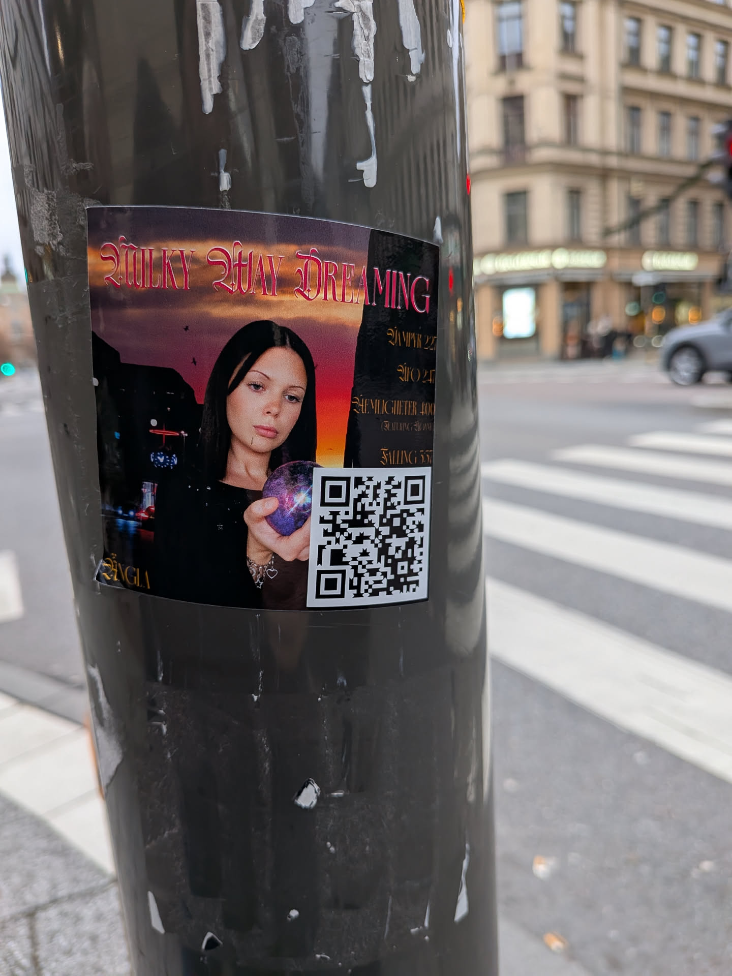 The sticker features a portrait of a young woman with dark hair, looking directly at the viewer. She is holding a glowing, purple and blue orb that resembles a miniature galaxy or nebula. The background is a stylized depiction of a night sky with a gradient of purple and orange hues, and silhouetted buildings and a few small birds. The text 'MILKY WAY DREAMING' is prominently displayed at the top in a decorative, pink font. Below the image, there is text that appears to be track listings or artist information, followed by a QR code.