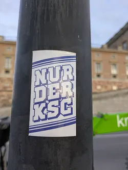 The sticker is rectangular with rounded corners and features bold, blue block letters spelling out 'NUR DER KSC' against a white background. The text is outlined in a darker blue. Two thin blue horizontal lines run across the top and bottom of the text, creating a border. The sticker is affixed to a dark, cylindrical object, likely a lamppost, with a blurred building and sky in the background.