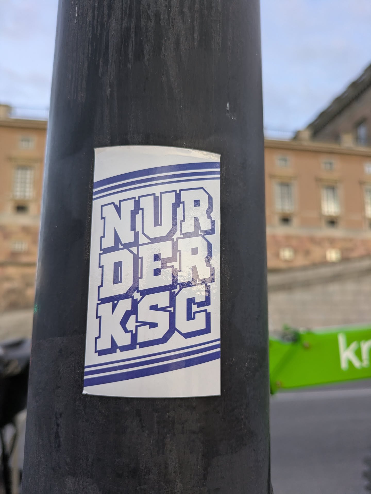 The sticker is rectangular with rounded corners and features bold, blue block letters spelling out 'NUR DER KSC' against a white background. The text is outlined in a darker blue. Two thin blue horizontal lines run across the top and bottom of the text, creating a border. The sticker is affixed to a dark, cylindrical object, likely a lamppost, with a blurred building and sky in the background.