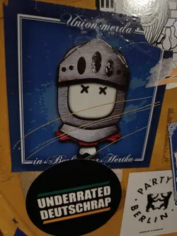 A sticker featuring a cartoon knight in a helmet with 'x' eyes. The background is blue with the text 'Union Merda' at the top and 'in Berlin Hertha' at the bottom. 