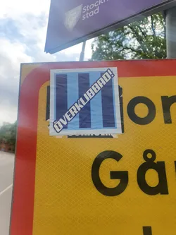 A sticker featuring vertical blue and white stripes with the text "ÖVERKLIBBAD!" diagonally across it. The sticker is affixed to a yellow traffic sign.