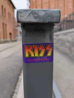 A sticker featuring the iconic KISS logo is affixed to a gray metal pole. Below the logo, smaller text reads '(Bandet inte drycken)', which translates to '(The band, not the drink)' in Swedish.